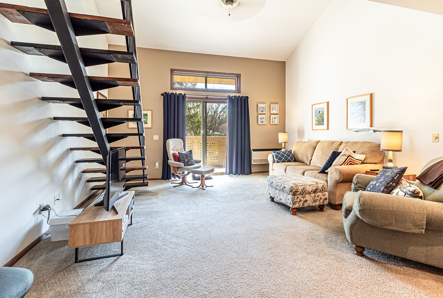 A brightly lit apartment living room with couch and chairs on the right, glass doors leading to a balcony, and stairs leading to a loft on the left