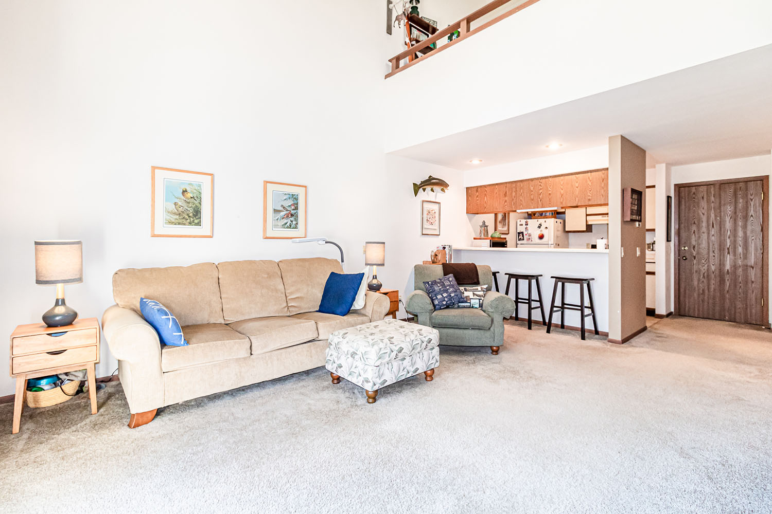 A brightly lit apartment living room with couches, a view of the galley kitchen and counter with stools in the background