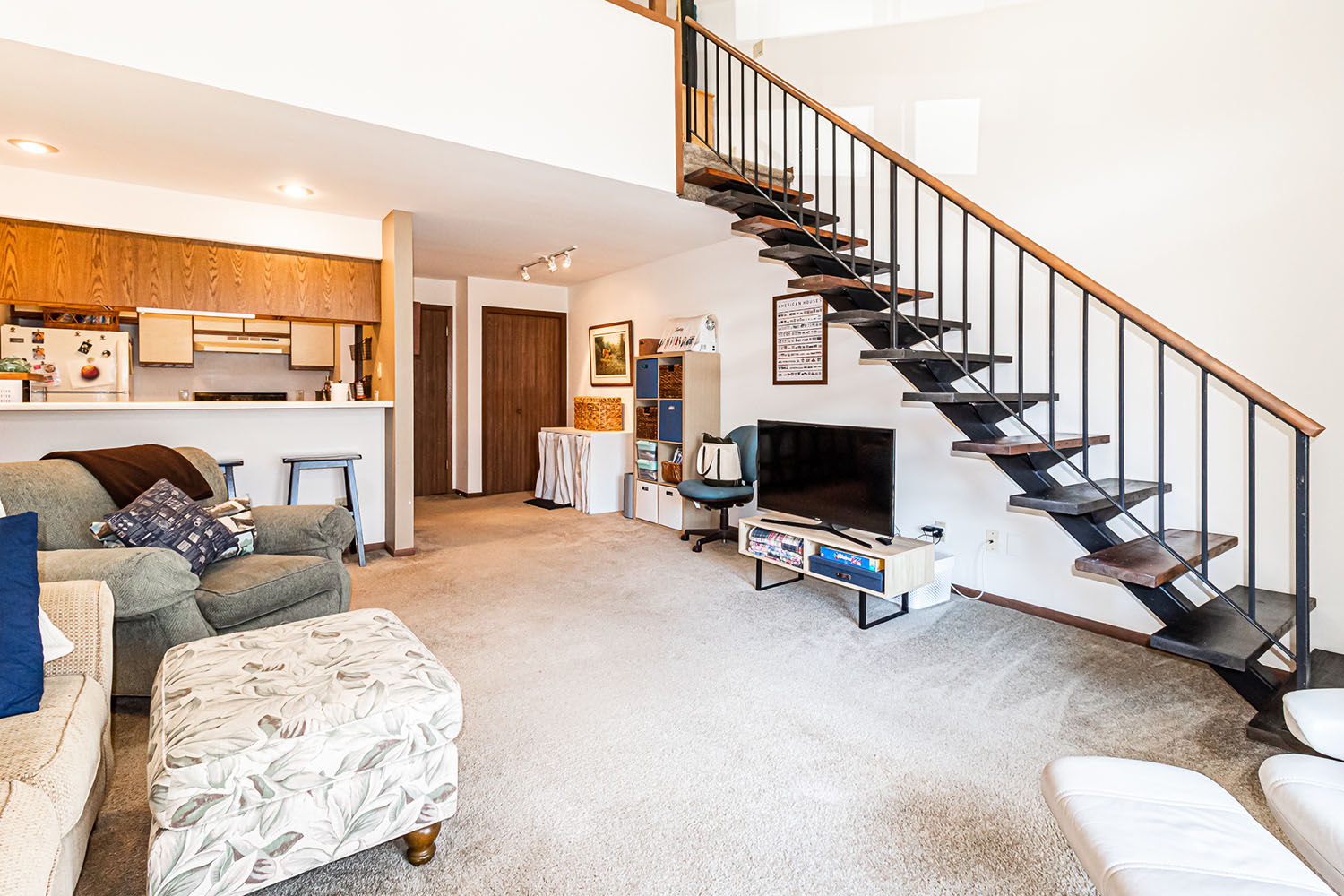 An apartment living room with couch and chairs, a tv and a view of the galley kitchen, stairs lead to a loft on the right