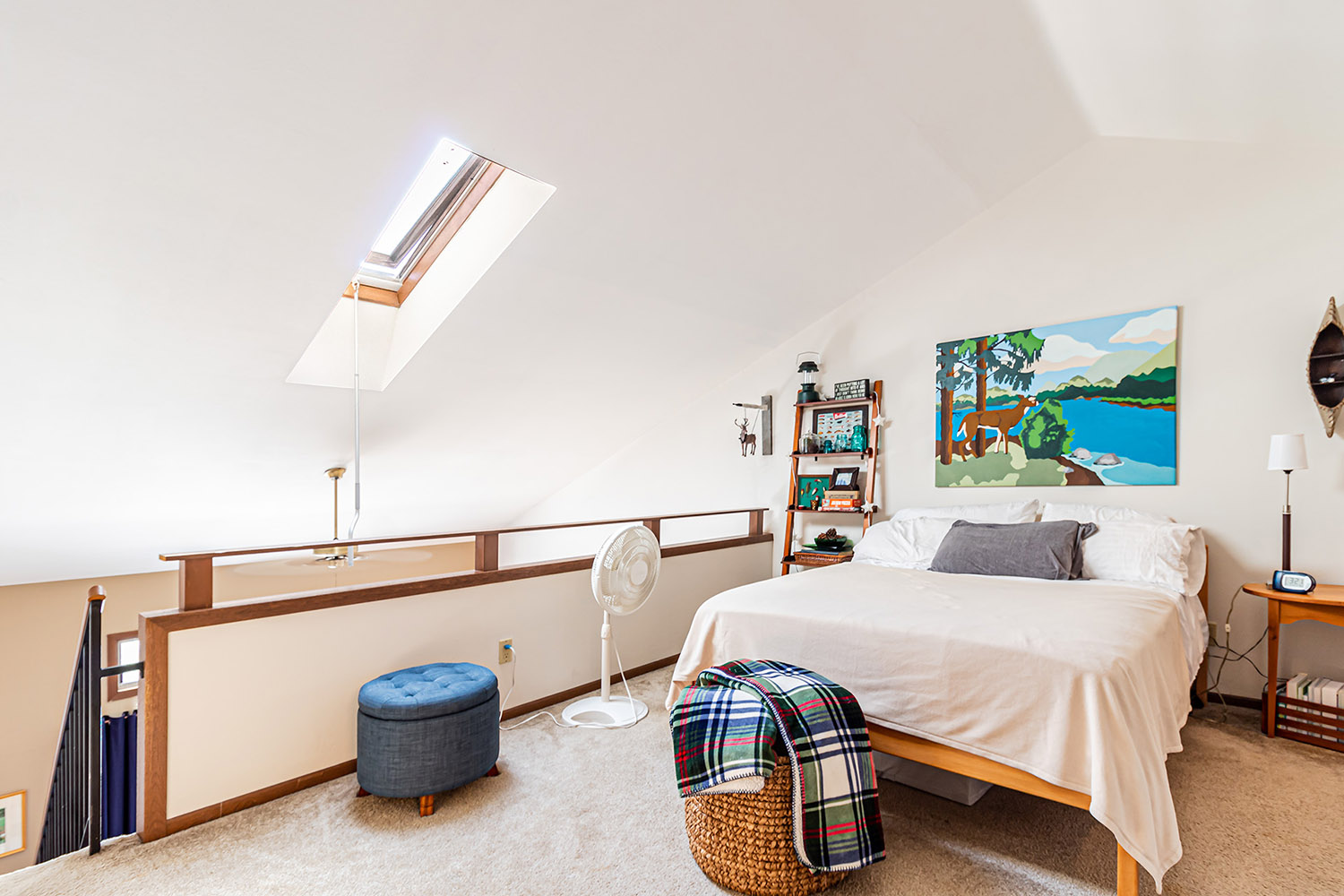 An apartment loft bedroom with a bed, bookshelf, side table, stool, fan, and a skylight