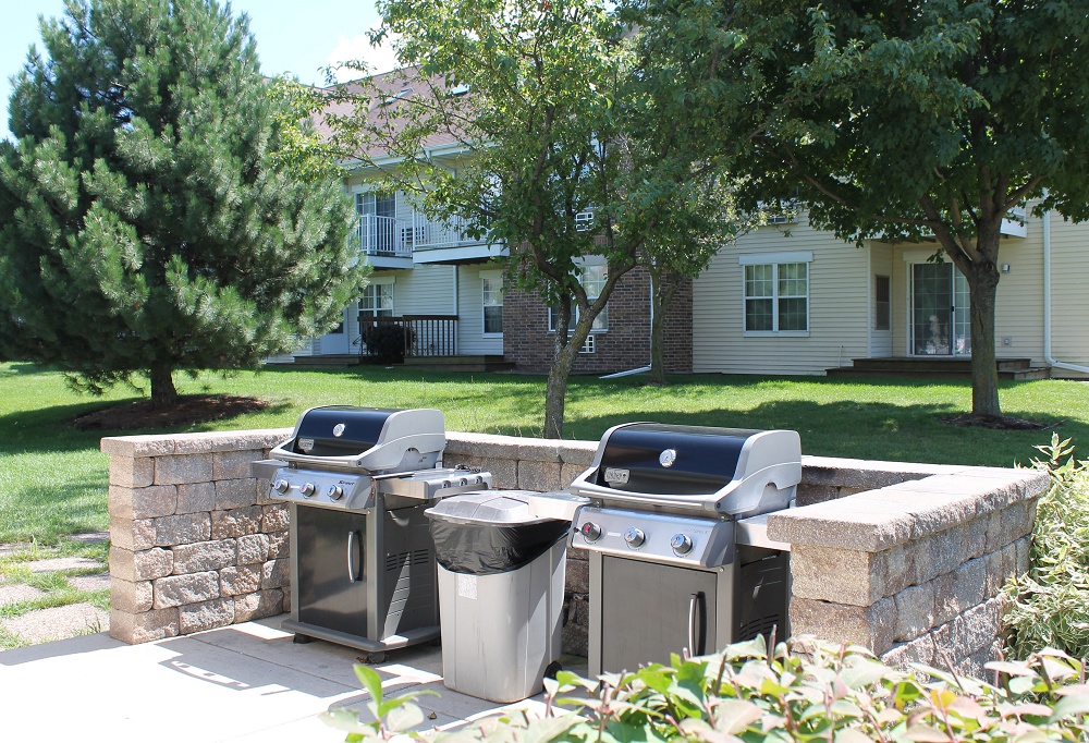 Two outdoor grills on a concrete patio with a short stone wall, and a lawn with trees leading up to the apartment complex