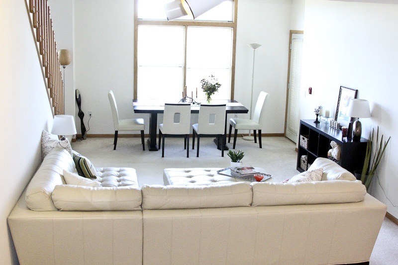 Apartment living room with large white couch, a tv on the right, a small table and chairs in front of a window, and stairs on the left