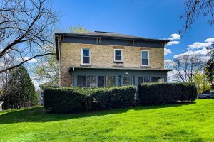 The front of a brick house with hedges in front and a grassy lawn on a sunny, early spring day