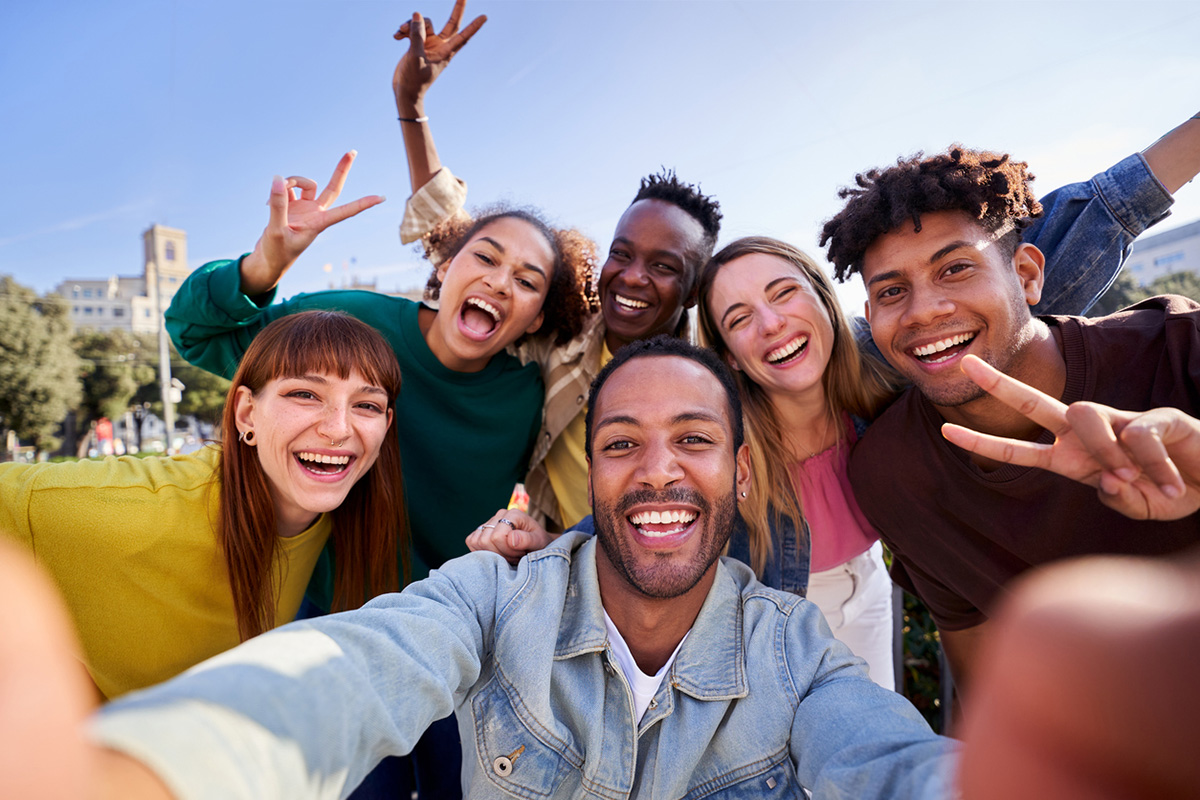 Happy multiethnic group of friends taking a selfie outdoors, having fun in sunny day. University students using phone app to take photos looking at camera.